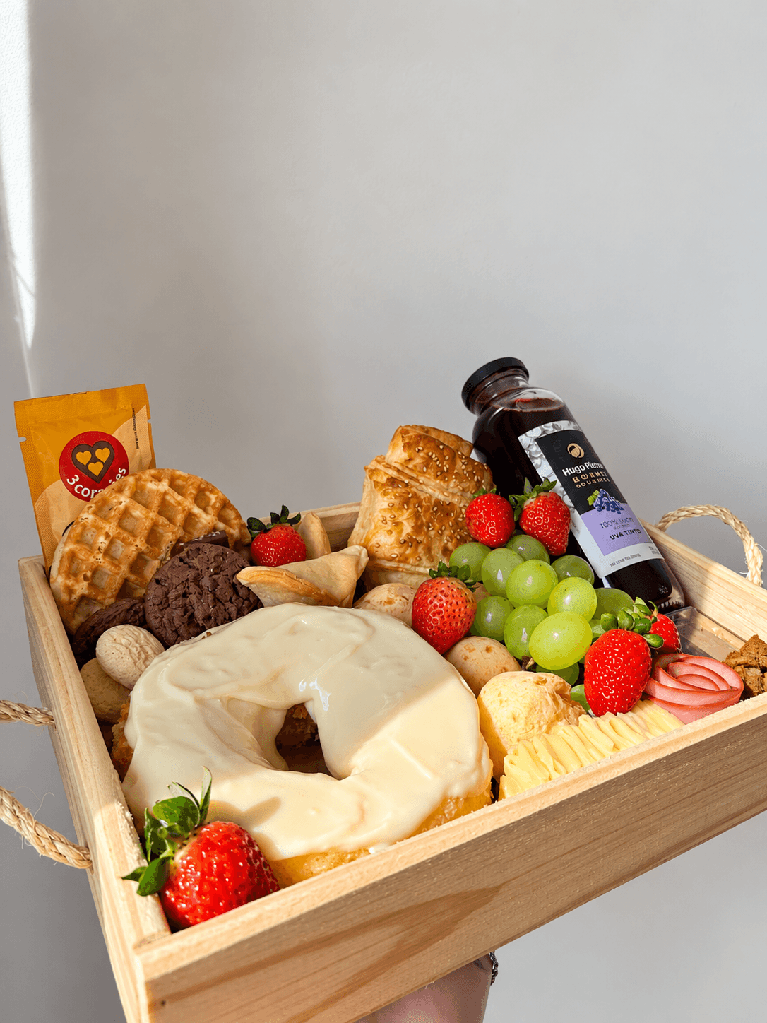 Cesta de madeira com bolo coberto de creme, waffle, cookies, morangos, uvas e uma garrafa de suco